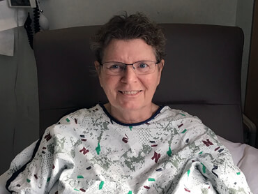 Vicki smiling and wearing a medical gown sits in a chair in her hospital room.