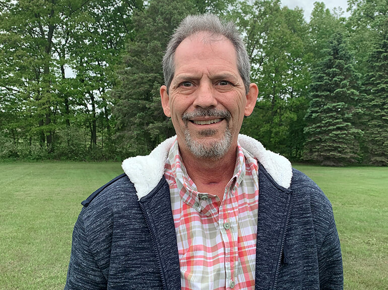 A smiling Kevin Gilkes stands outside in front of a bank of evergreen trees.